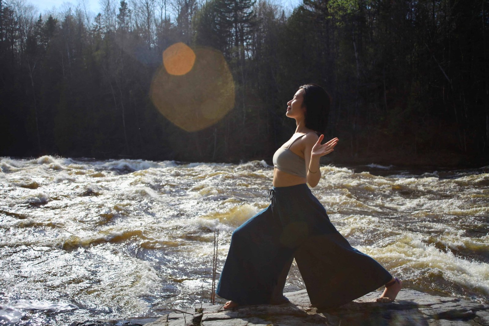 Sao Aum faisant du Qi Gong sur des rochers près d'une rivière et d'une forêt, avec les bras au niveau des épaules.