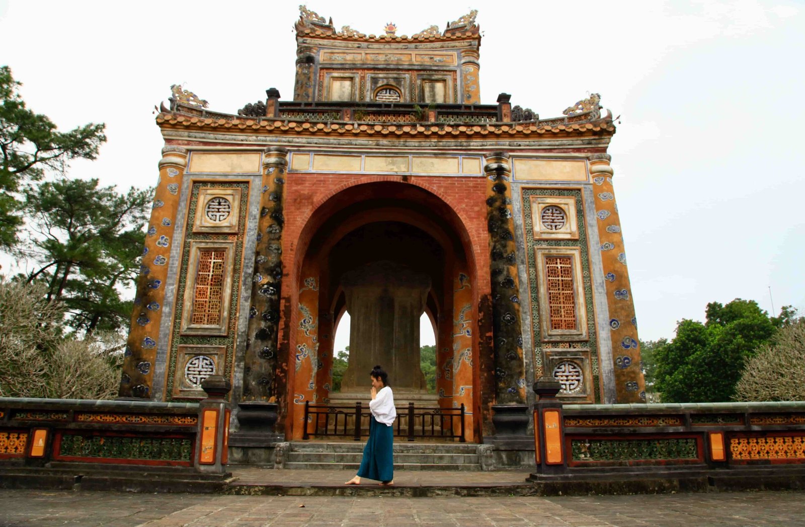 Sao Aum, au loin, en posture de prière, de côté,nu pieds, avec derrière elle une arche vietnamienne.